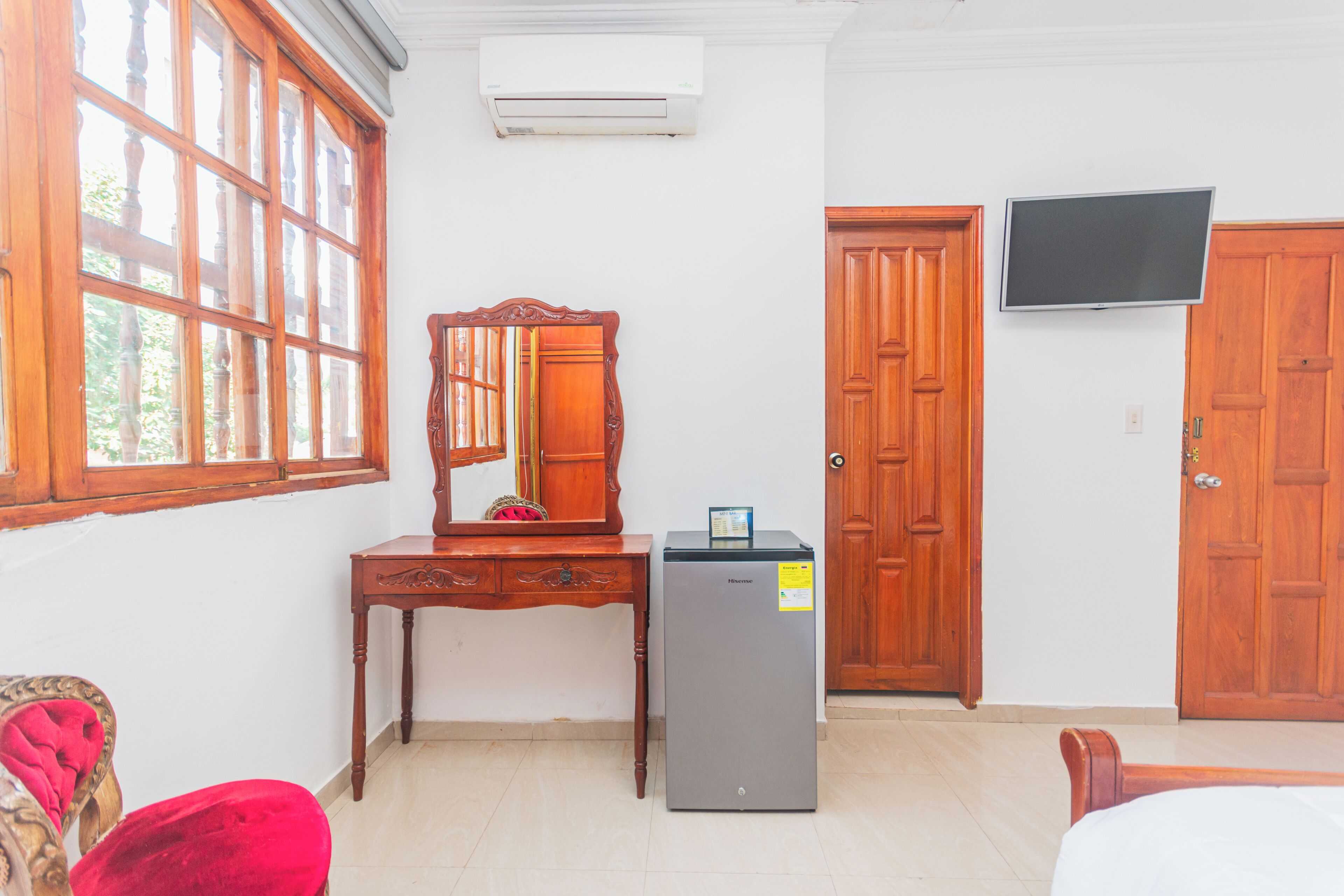 Una habitación con un escritorio de madera, un refrigerador pequeño, un televisor montado en la pared y un ventanal con marcos de madera.