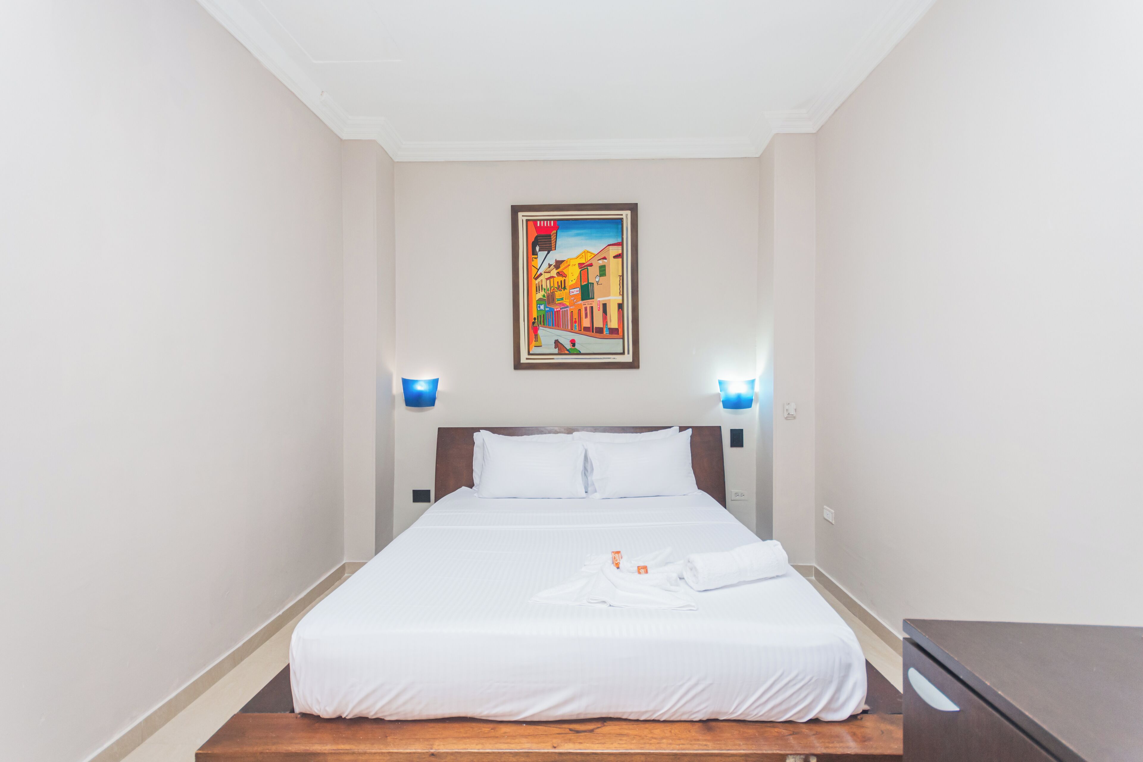 A hotel room with a bed, a wooden bench, a painting on the wall, and two wall-mounted lamps.