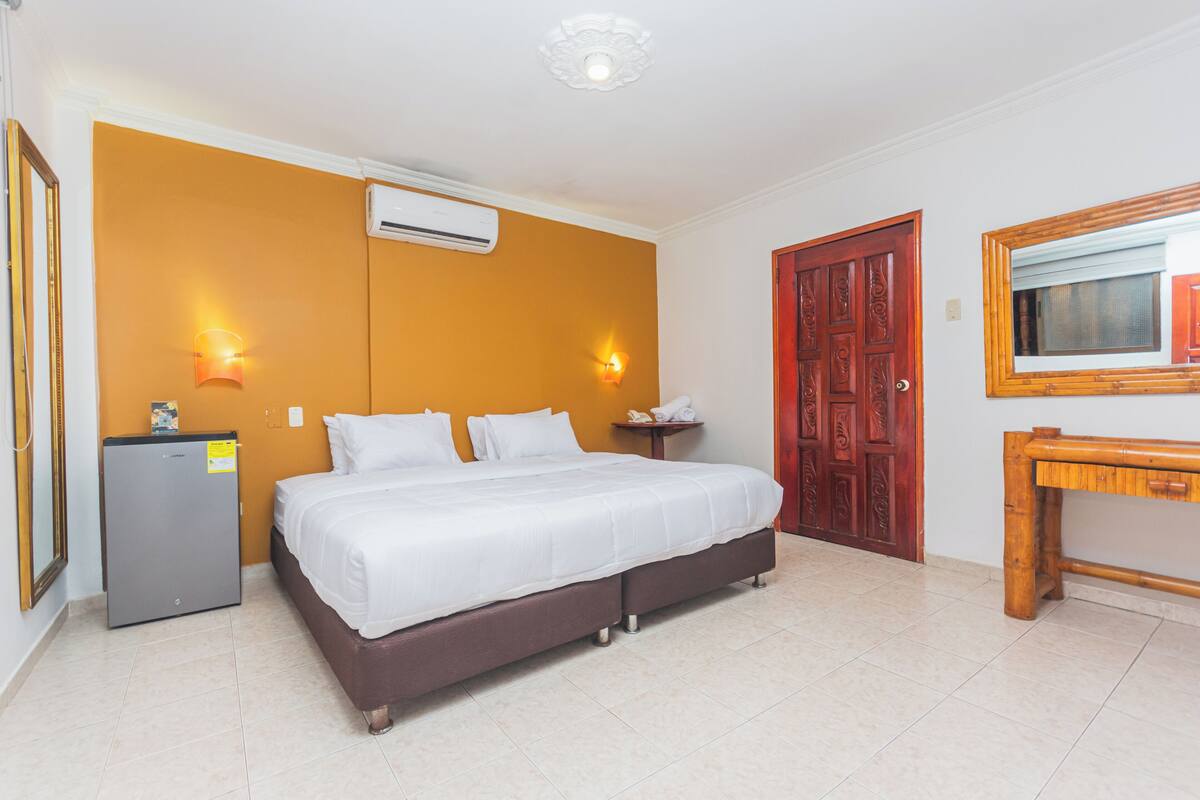 A hotel room with a bed, a desk, a mirror, and an air conditioning unit.