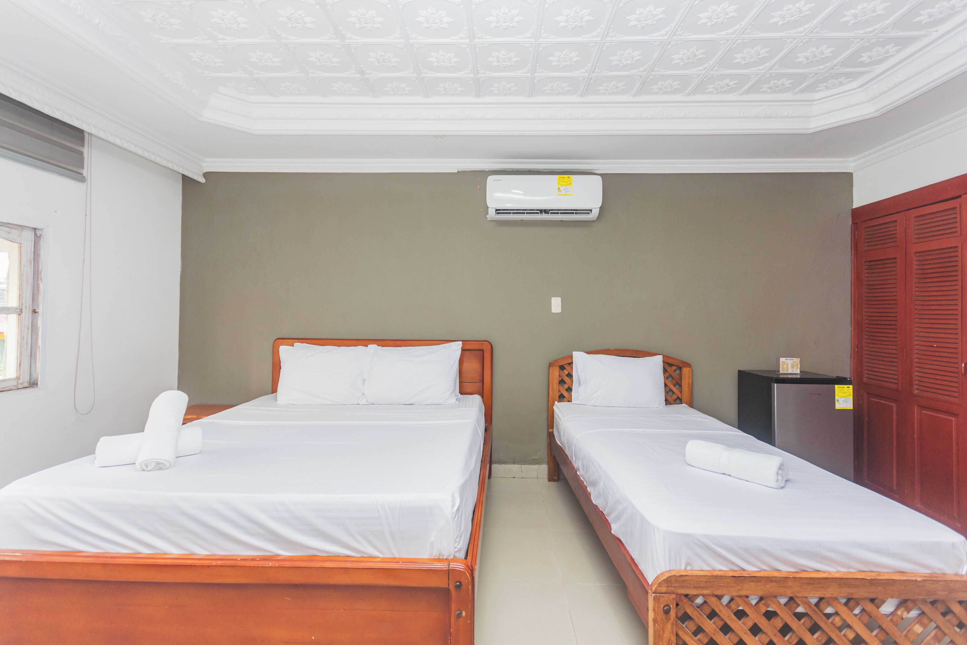 A hotel room with two wooden beds, white linens, an air conditioning unit, a small black refrigerator, and a red door.