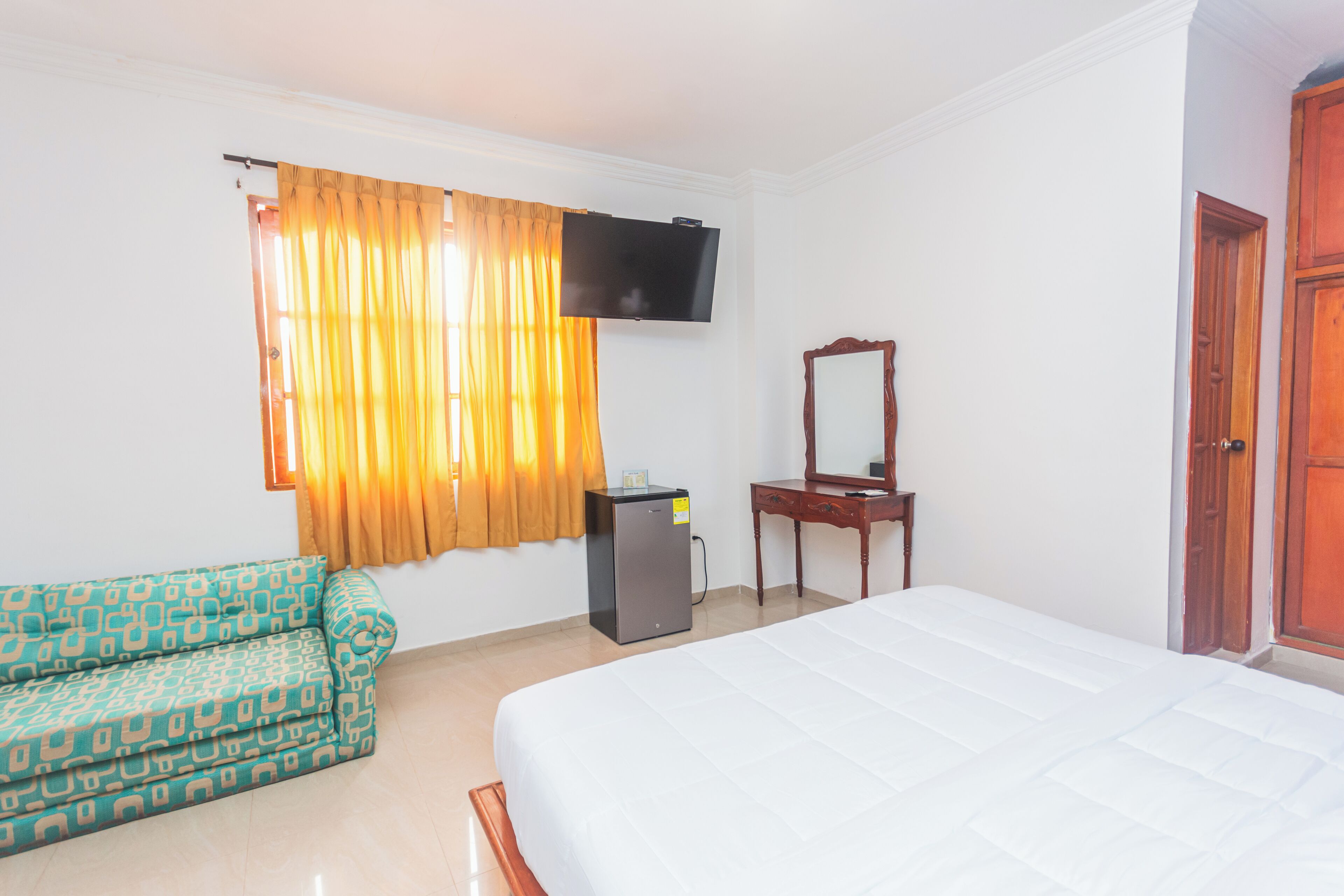Een hotelkamer met een bed, een slaapbank, een aan de muur bevestigde televisie, een kleine koelkast en een houten deur.