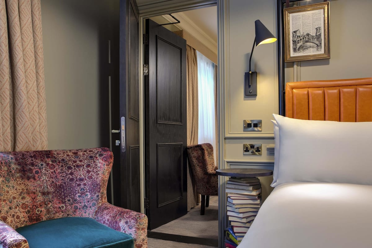Een hotelkamer met een bed, een stoel, een bureau met lamp en een boekenplank.