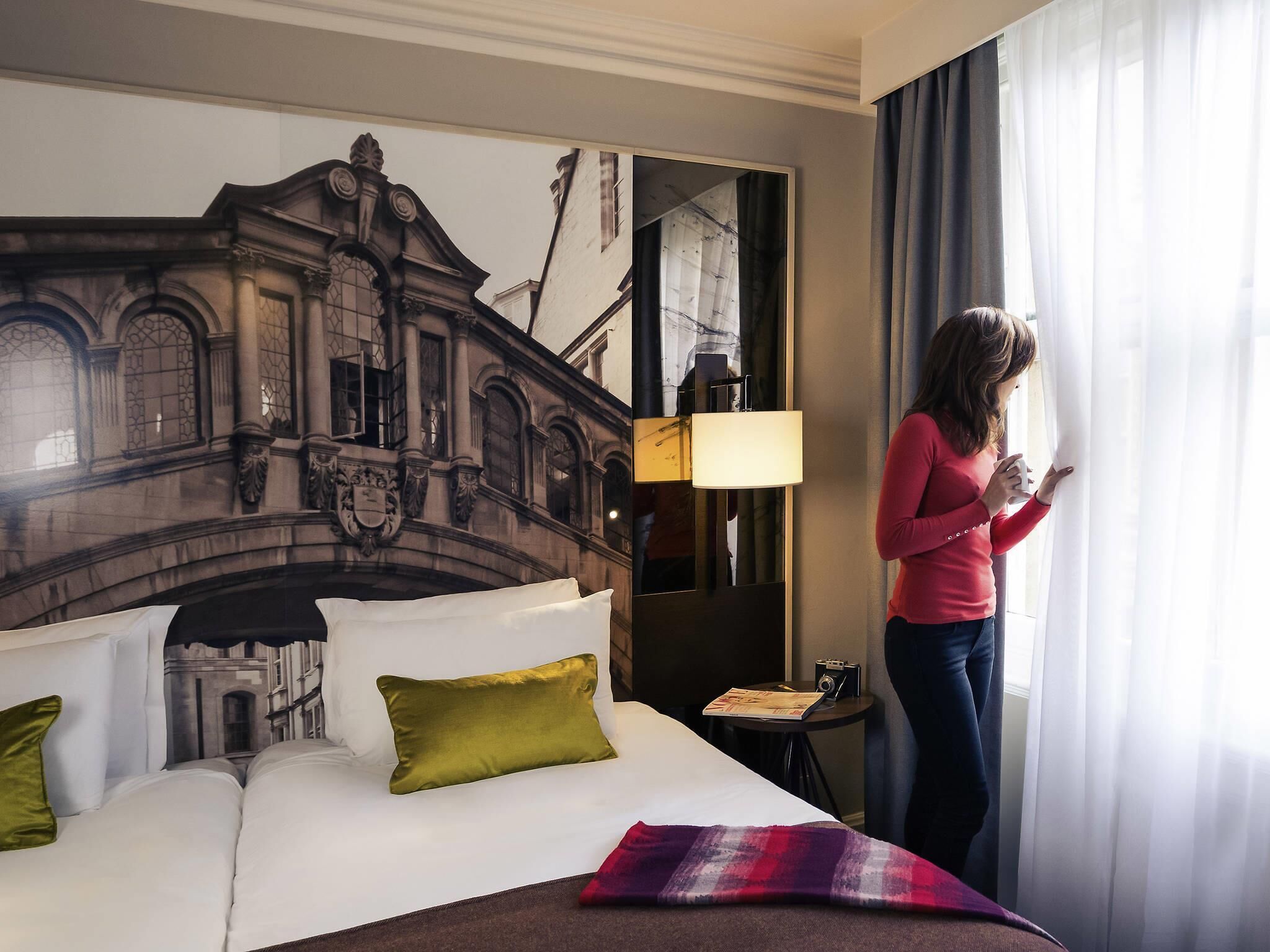 Een hotelkamer met een bed, kussens, een lamp, een tafeltje met een tijdschrift en een raam met gordijnen.