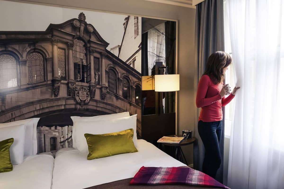 A hotel room with a bed, pillows, a lamp, a table with a magazine, and a window with curtains.