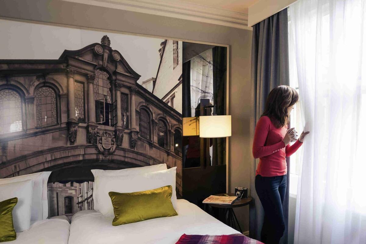 Een hotelkamer met een bed, kussens, een lamp, een nachtkastje en een groot raam met gordijnen.