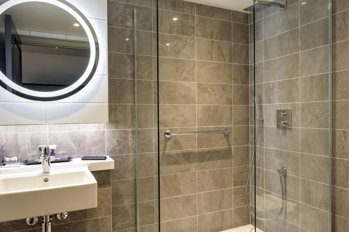 A modern bathroom with a glass-enclosed shower, a white sink with chrome fixtures, and a large mirror with a circular light.