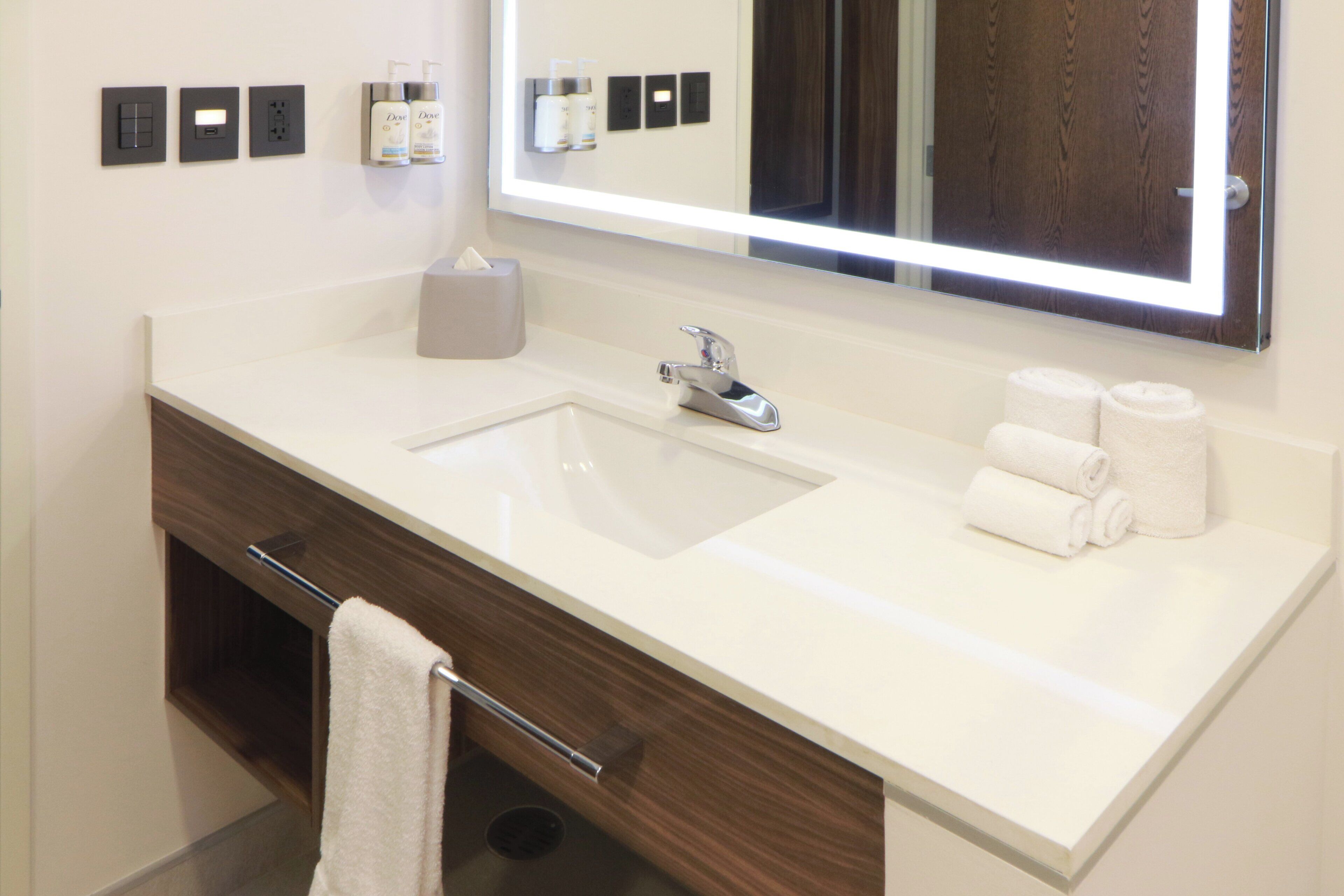 Un baño moderno con un lavamanos blanco, un espejo con iluminación integrada, un perchero y artículos de aseo en la pared.