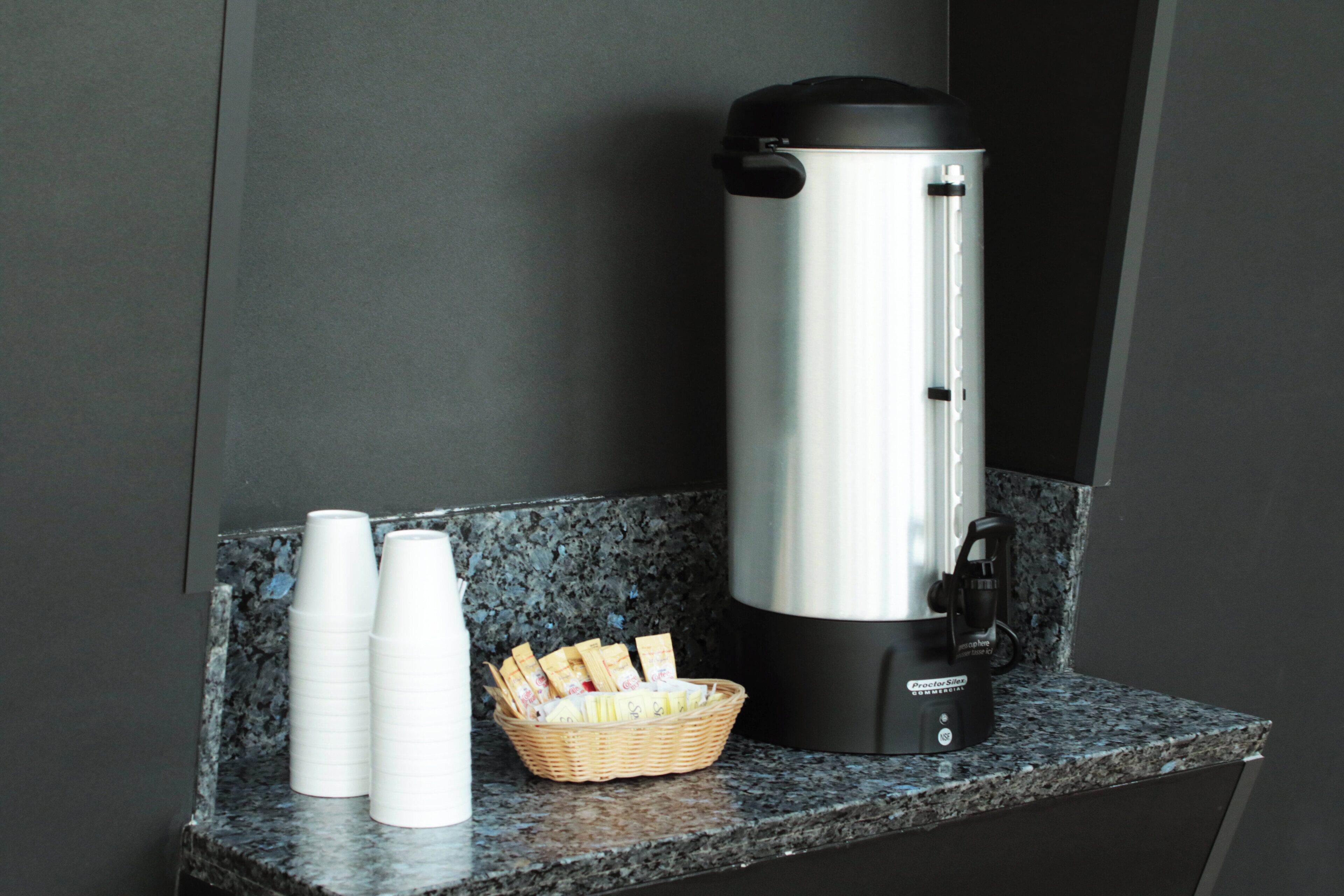 A stainless steel water dispenser on a granite countertop, a basket of snacks, and two white paper cups.