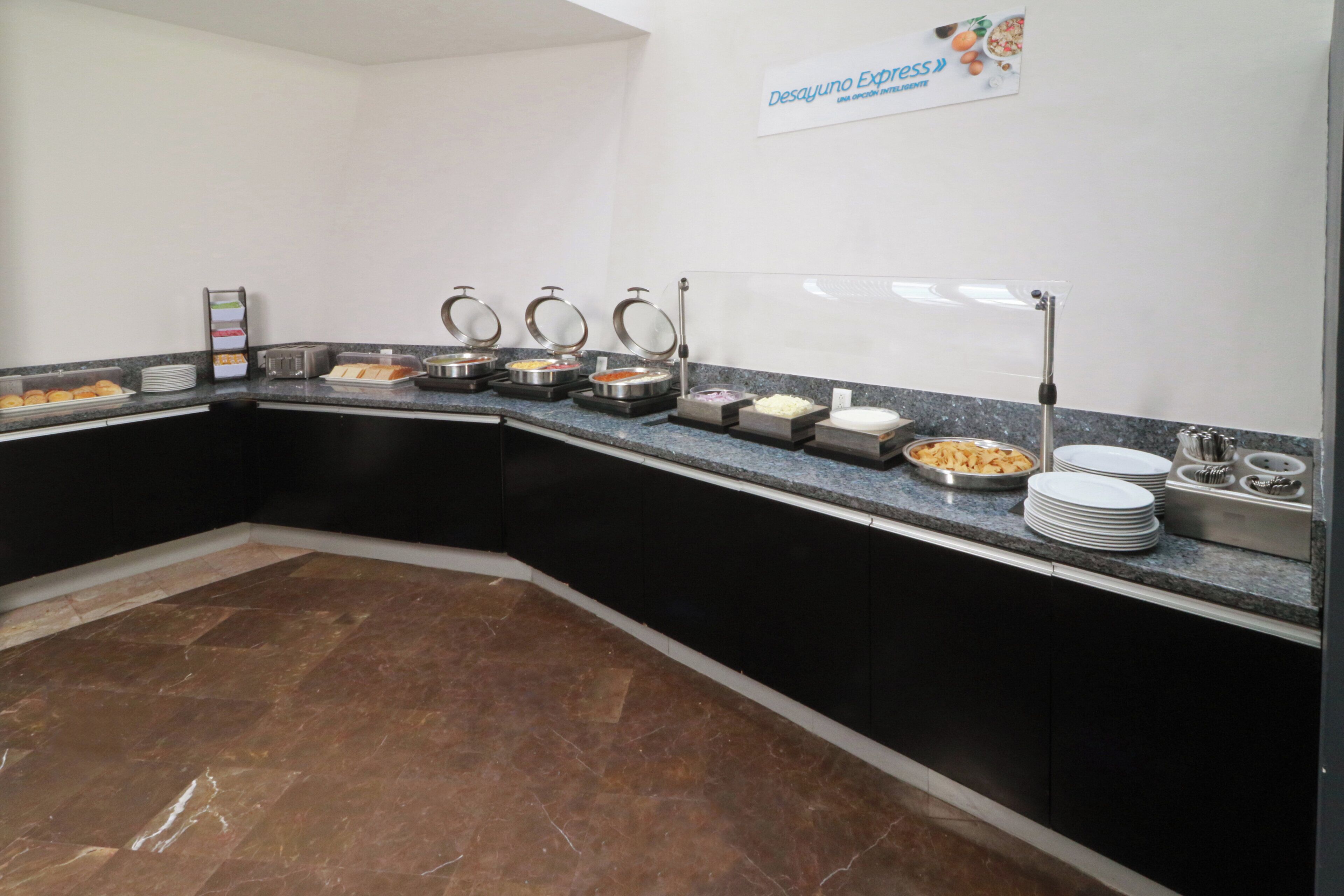 A buffet setup with various food containers, a sign for Desayuno Express, and a marble-tiled floor.