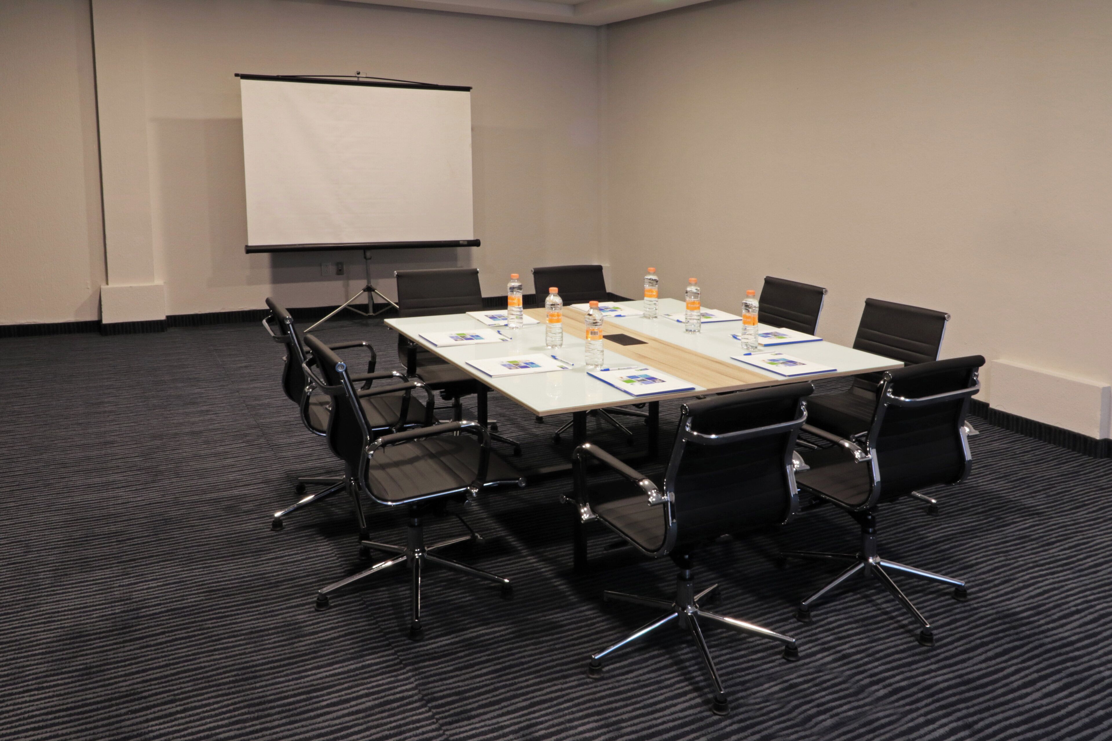 A conference room with a whiteboard, a projector screen, a table with chairs, water bottles, and documents.
