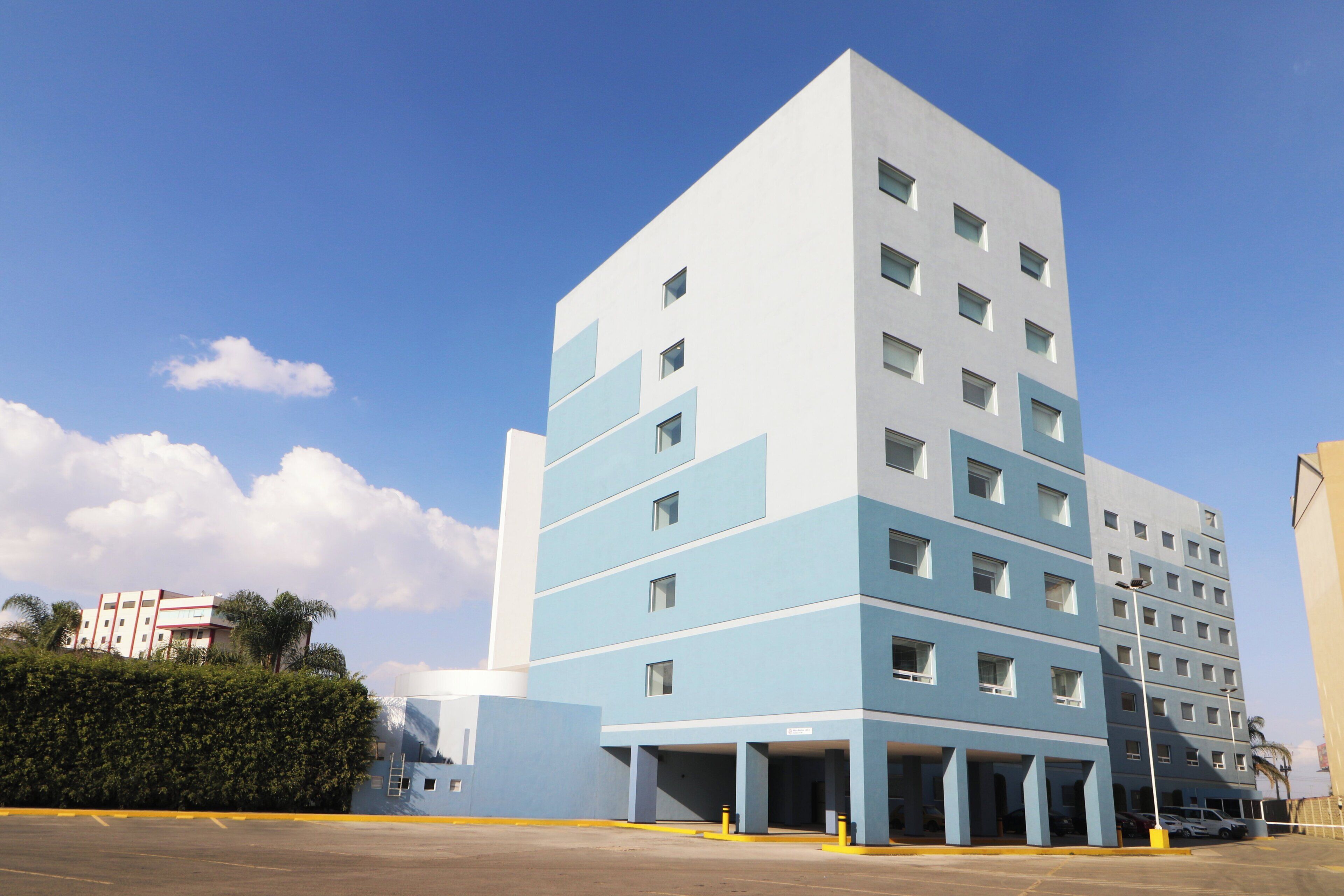 A multi-story building with a blue and white facade, multiple windows, and a parking area in front.