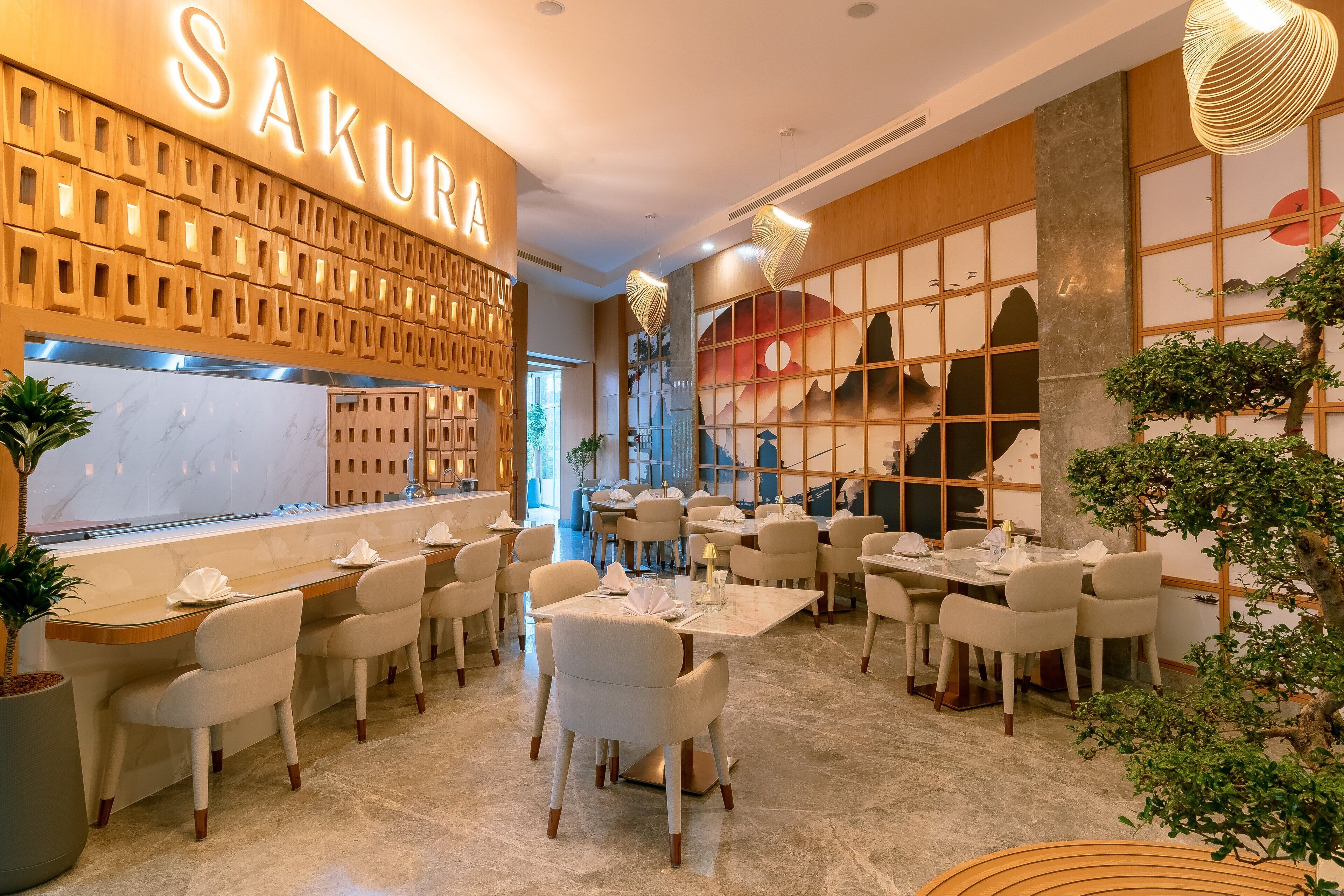Interior de un restaurante con una pared de madera que muestra 'SAKURA' en letras iluminadas, varias mesas con sillas y un amplio ventanal con vistas a una montaña.