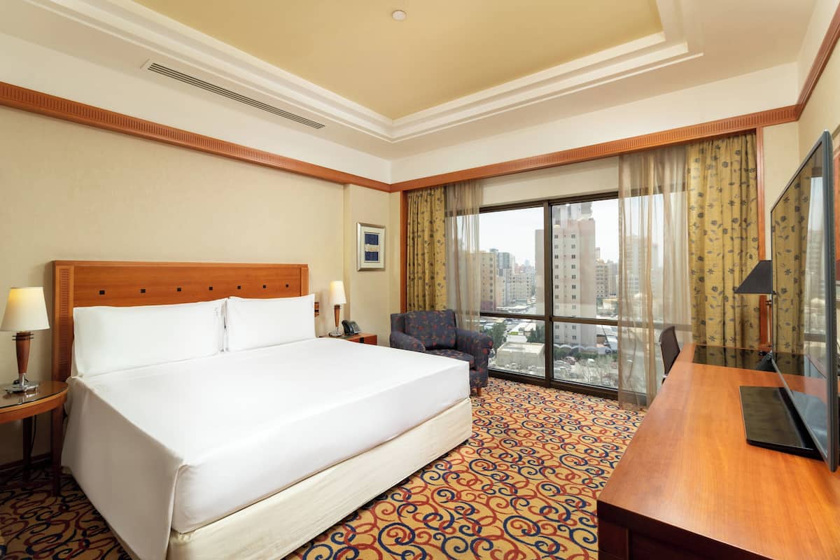 A hotel room with a large bed, a desk, a chair, and a large window offering a city view.