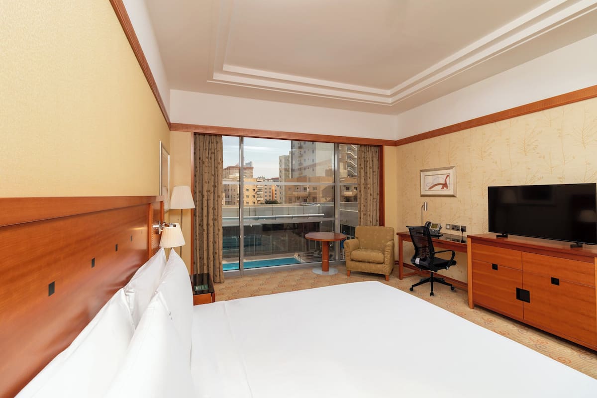 A hotel room with a large bed, a desk with a chair, a television, and a view of the city through large windows.