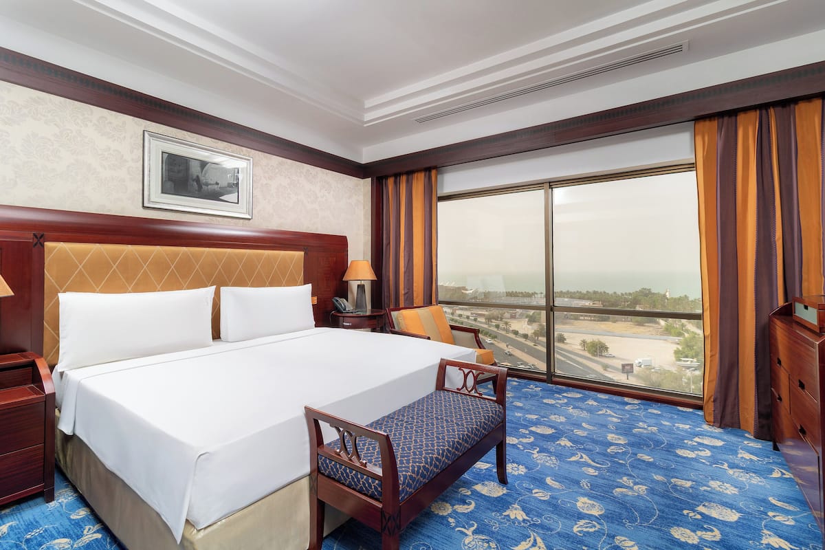 A hotel room with a large bed, a wooden headboard, a window with curtains, a nightstand, a lamp, and a view of the cityscape.