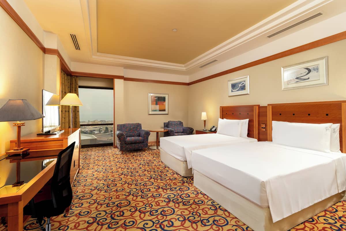 A hotel room with two beds, a desk, a chair, and a view of the city.