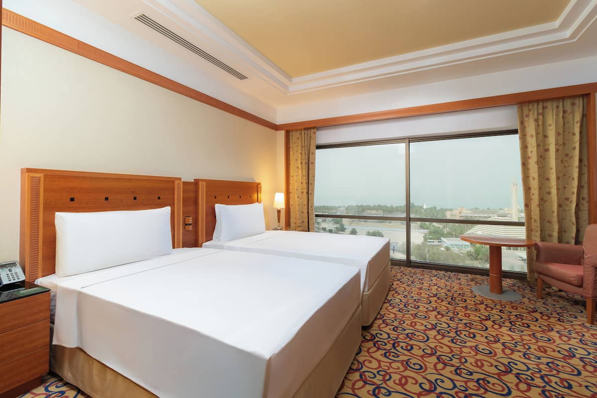 A hotel room with a large bed, a small table, a chair, and a view of the city through a large window.
