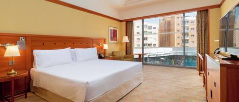 Habitación de hotel con una cama grande, un escritorio con lámpara, una silla, un televisor y vistas a la ciudad a través del ventanal.