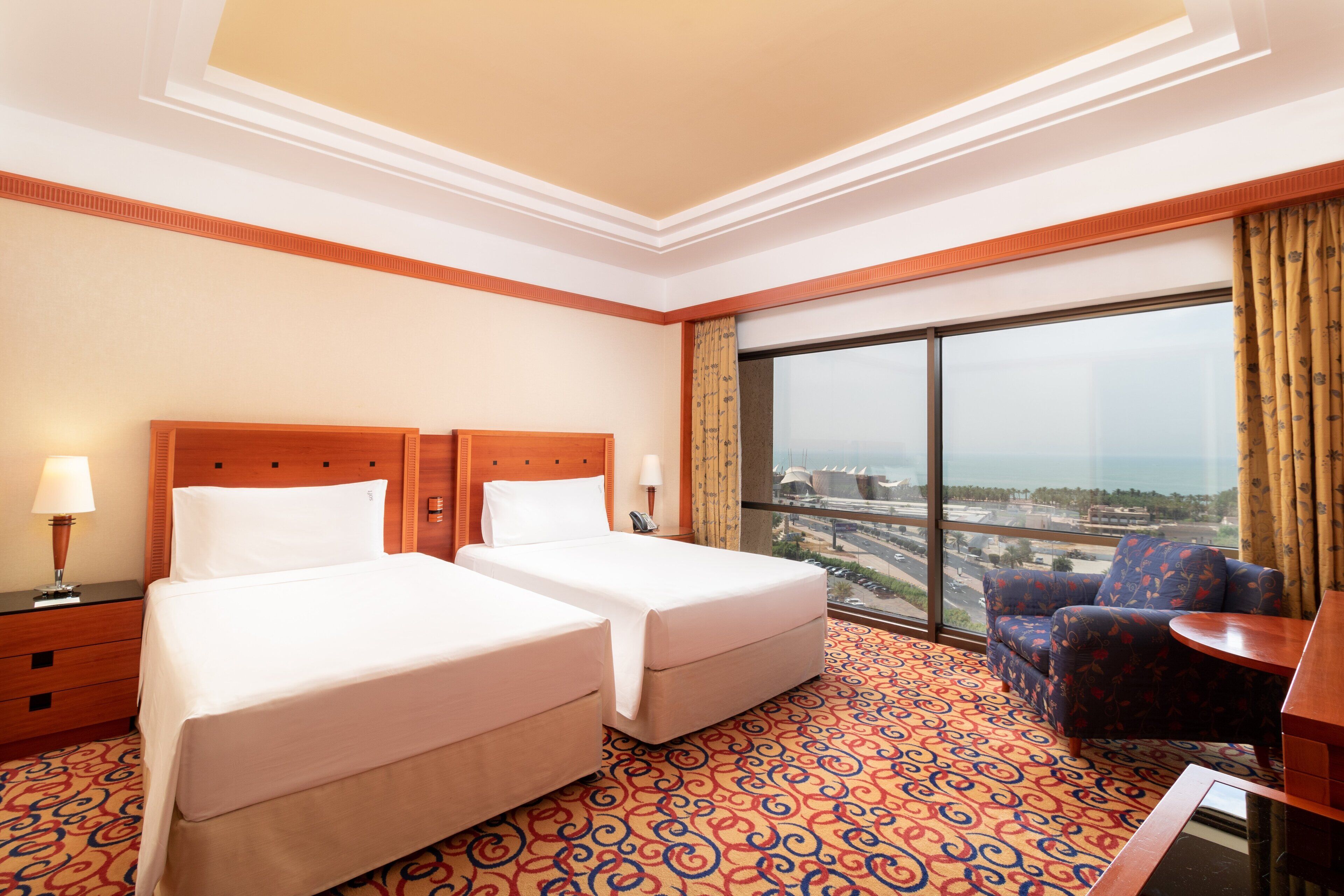 A hotel room with two beds, a large window with a city view, a patterned carpet, and a small table.