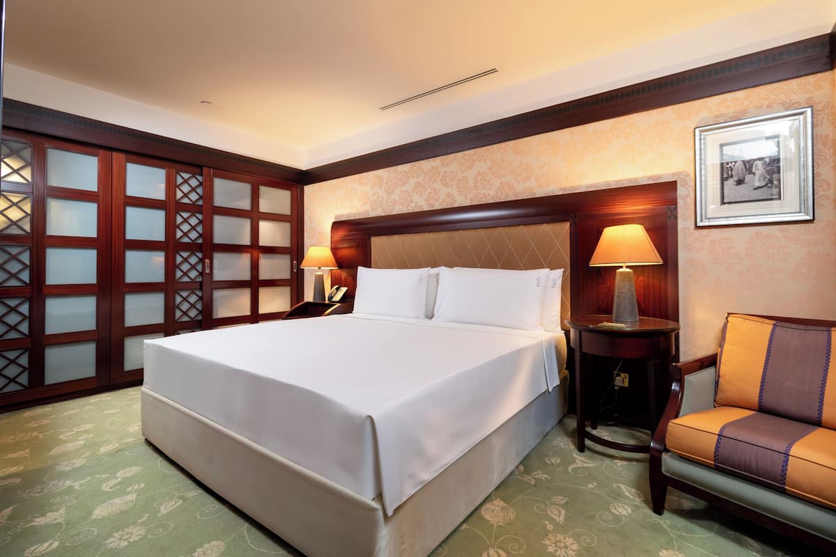 A hotel room with a large bed, two bedside tables with lamps, a chair, and a decorative wall panel.