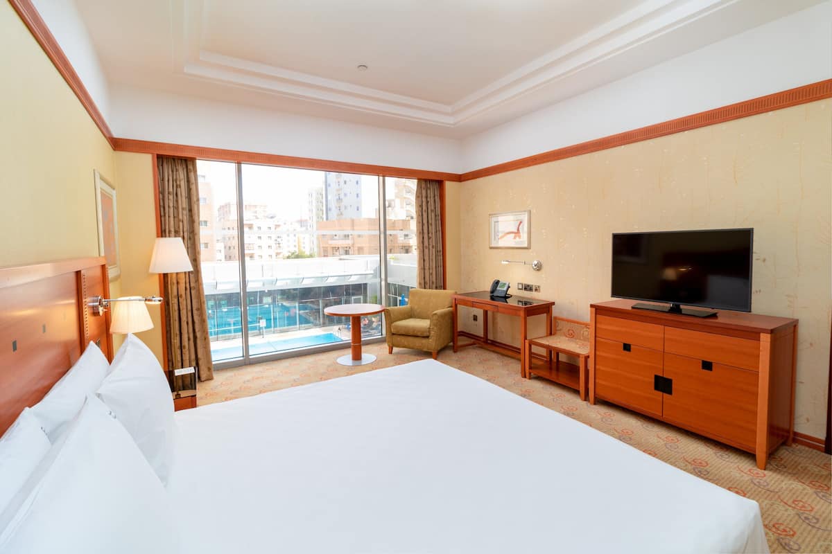 A hotel room with a large bed, a desk with a chair, a television, and a view of the city through large windows.