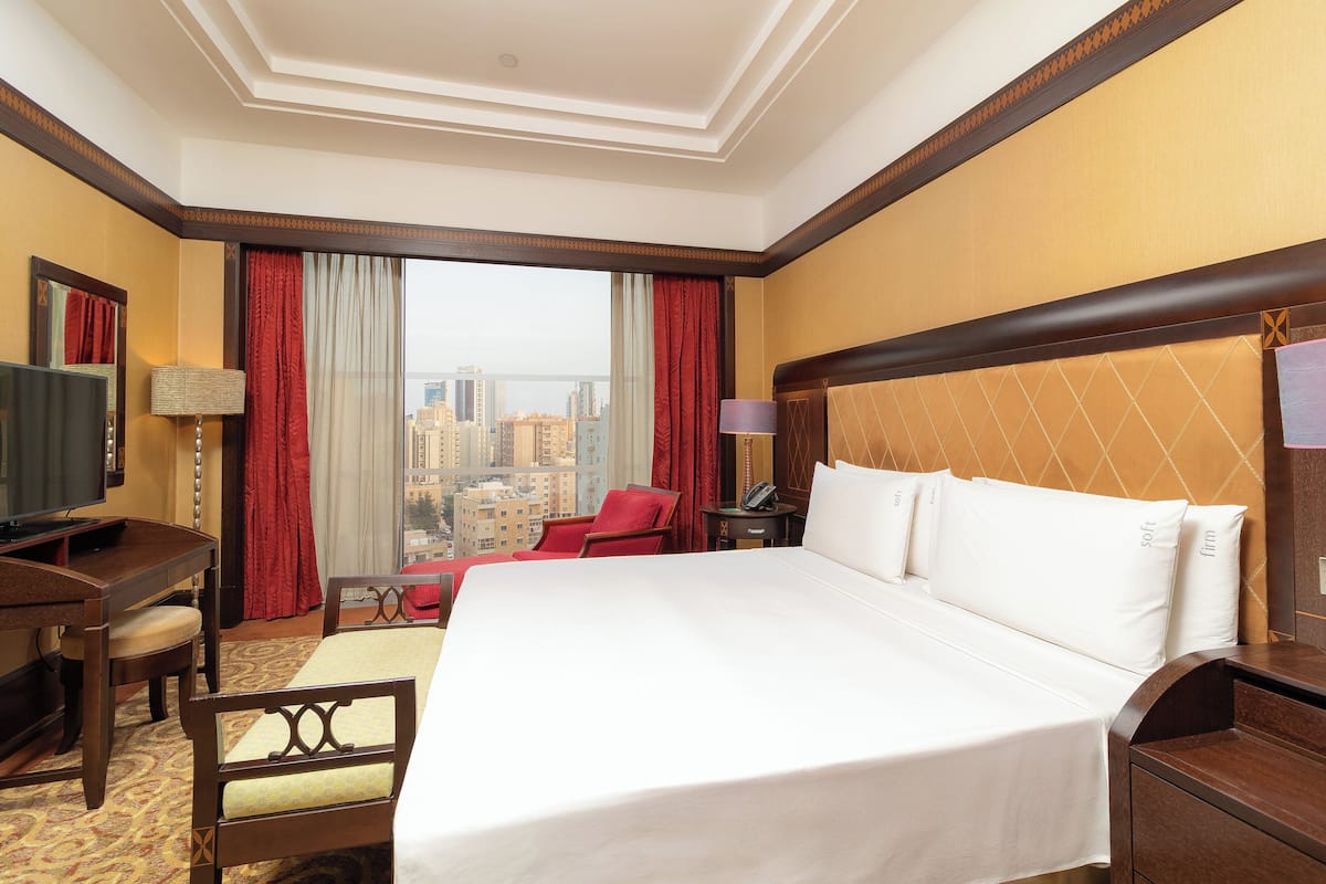 A hotel room with a large bed, a desk, a chair, a TV, and a view of the cityscape.