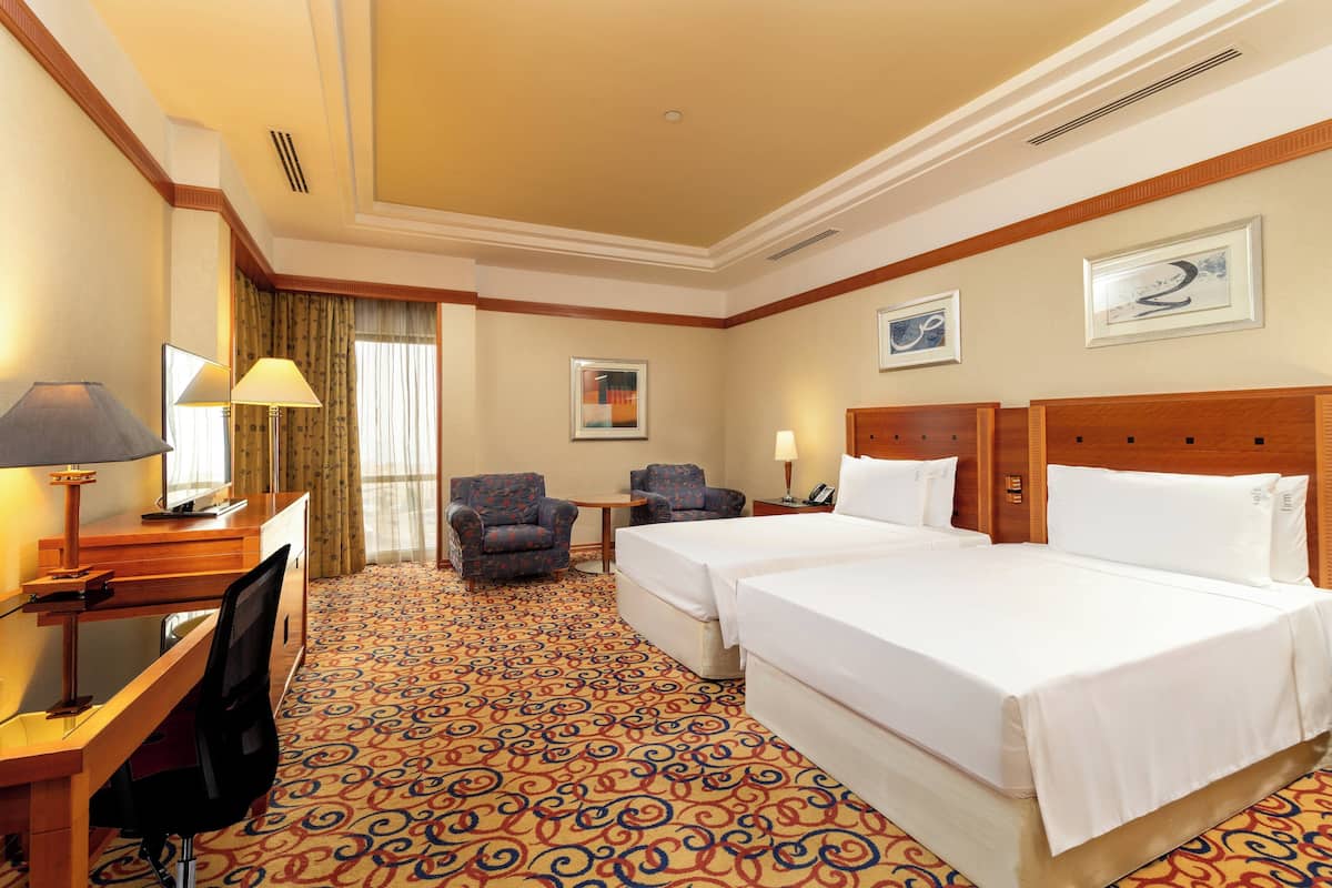 A hotel room with two beds, a desk with a chair, a small table, and a lamp.