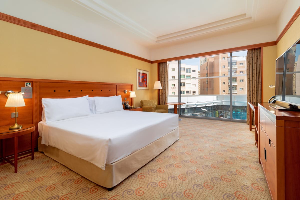 A hotel room with a large bed, two bedside lamps, a desk with a lamp, a chair, a TV, and a view of the city through large windows.