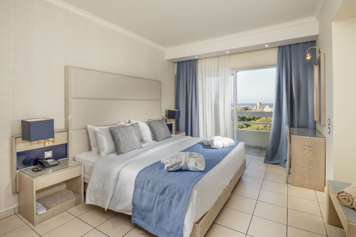 A hotel room with a large bed, a bedside table with a phone, a view of the city through the window, and a balcony with a view.