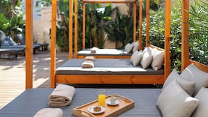 A wooden pergola with a cushioned seating area, a tray with a beverage and a snack, and a towel on the seat. - Athineon Hotel (Rhodes)
