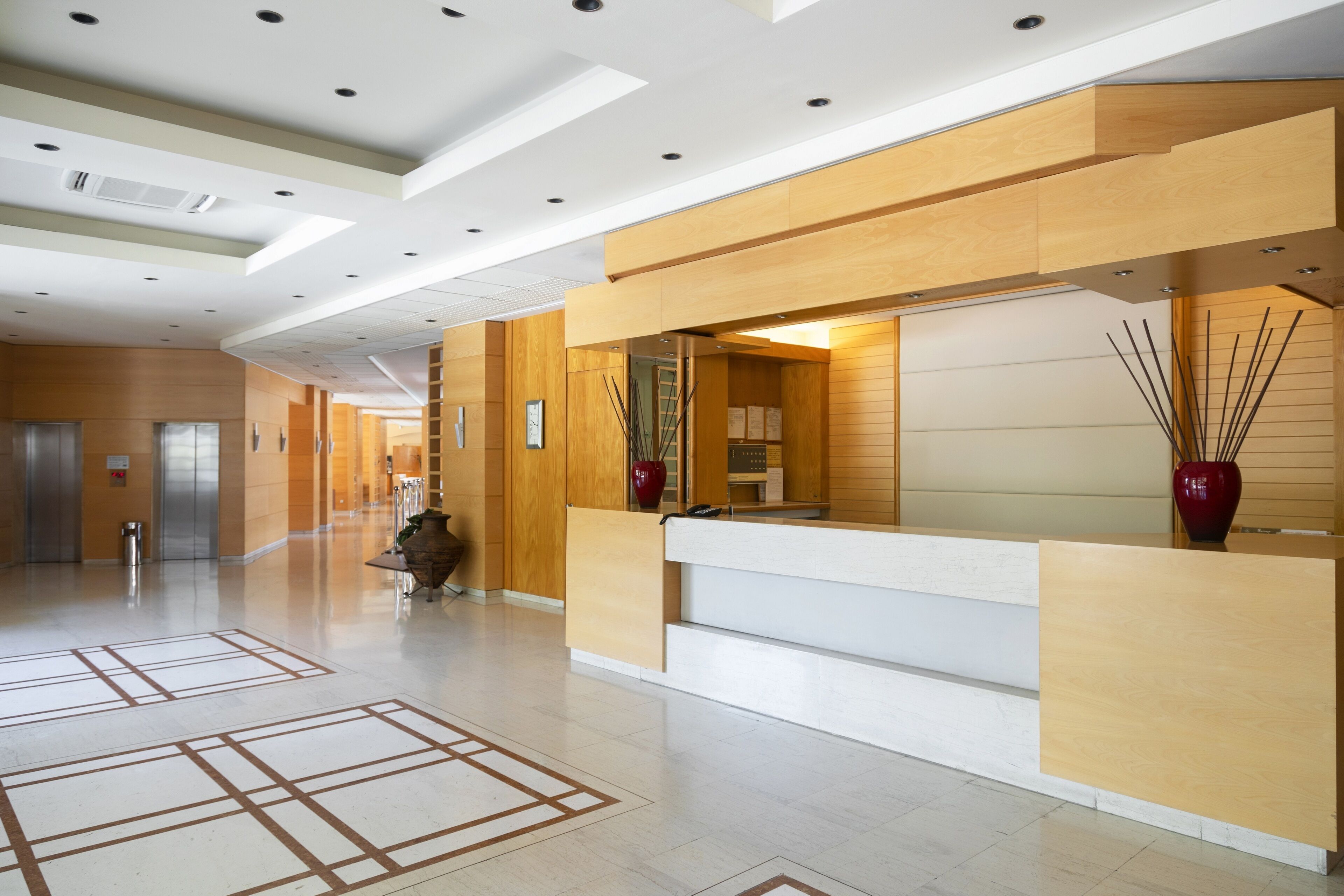 Egy modern szállodai előcsarnok, amelyben recepció, lépcsőház és egy nagyméretű váza található.