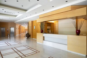 Egy modern szállodai előcsarnok, amelyben recepció, lépcsőház és egy nagyméretű váza található.