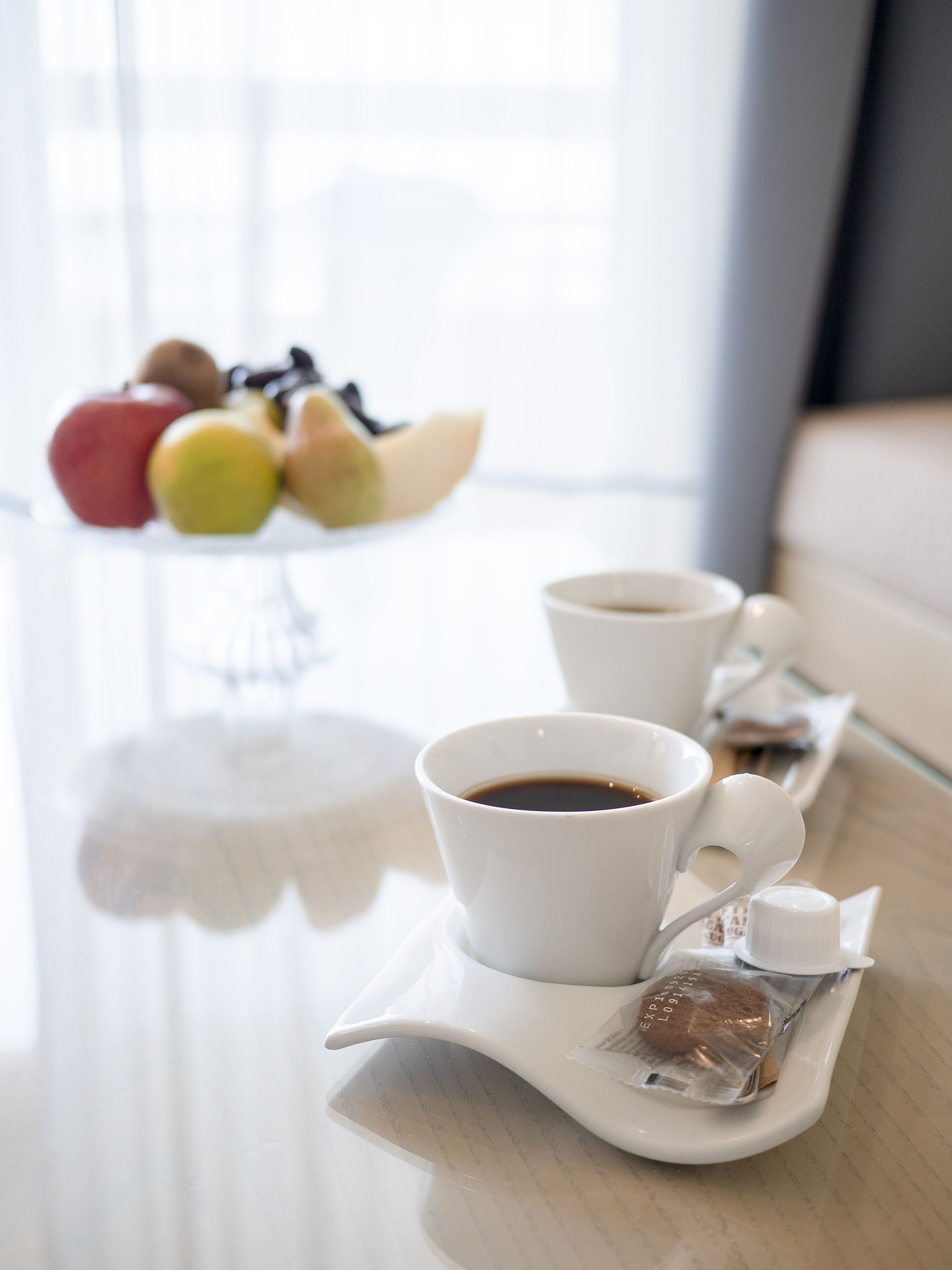Ein Frühstückstisch mit zwei Kaffeetassen, einer Keksplatte und einer Obstschale im Hintergrund.