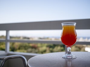 A glass of red and orange drink on a table with a view of a balcony and greenery in the background. - Athineon Hotel (Rhodes)