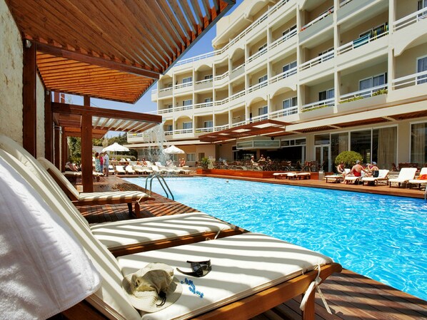 A hotel pool area with sun loungers, a wooden deck, and a building with multiple balconies. - Athineon Hotel (Rhodes)