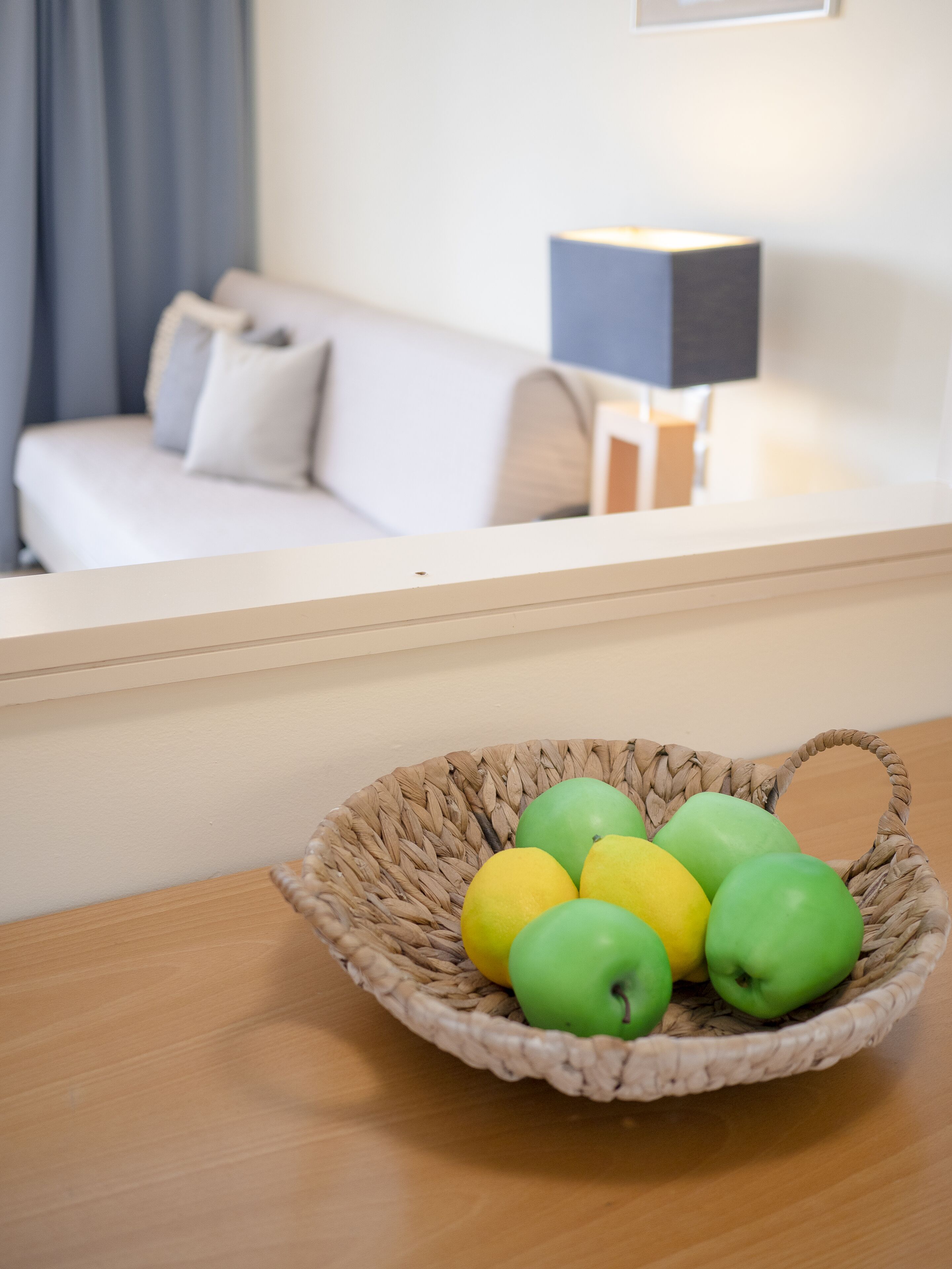 Un panier en osier contenant des pommes vertes et jaunes, posé sur une table en bois dans une pièce où se trouvent un canapé blanc et une lampe.
