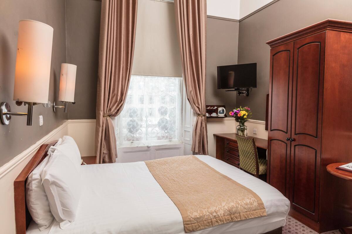 A hotel room with a bed, curtains, a TV, a wardrobe, and a desk with a vase of flowers.
