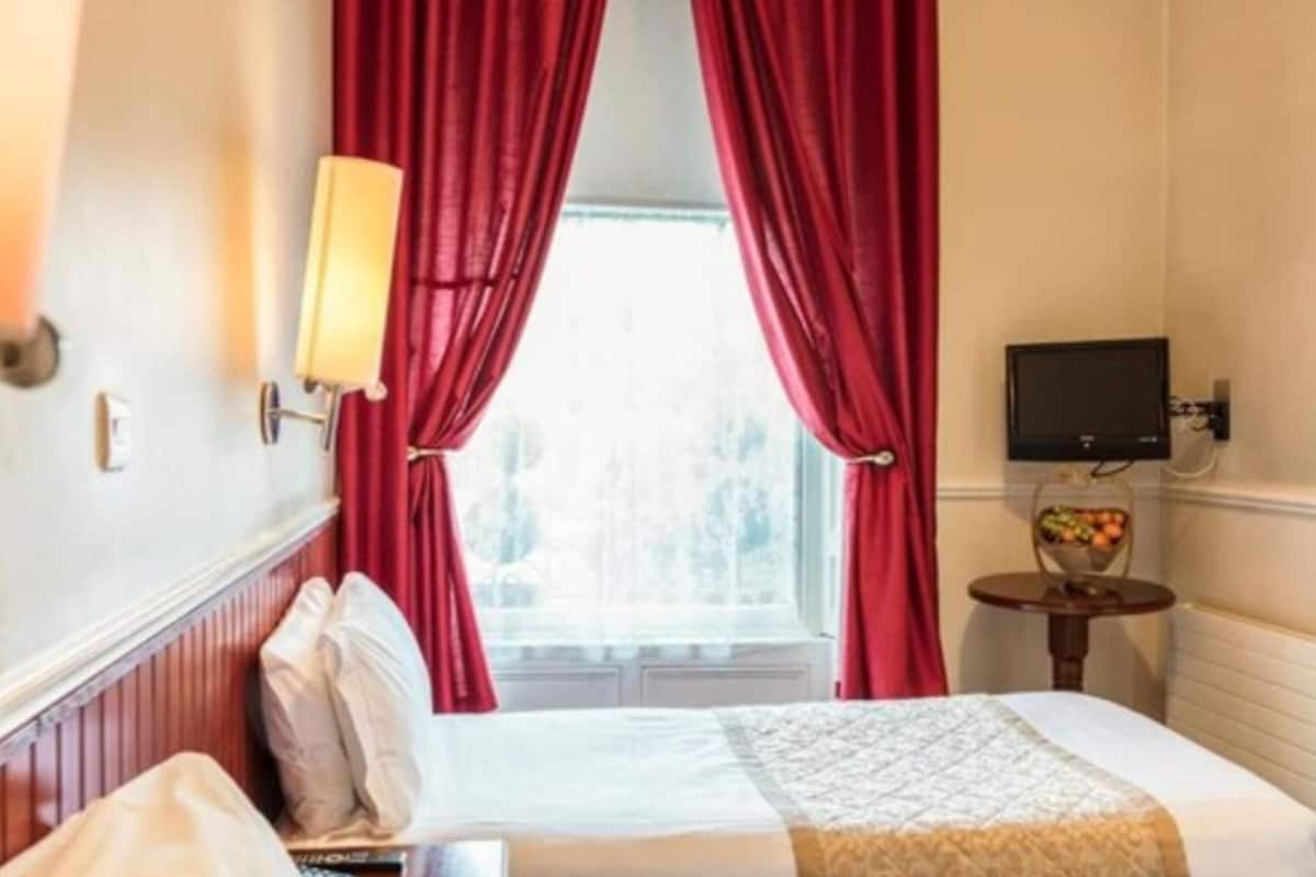 A hotel room with a bed, red curtains, a bedside lamp, a television, and a small table with fruit.