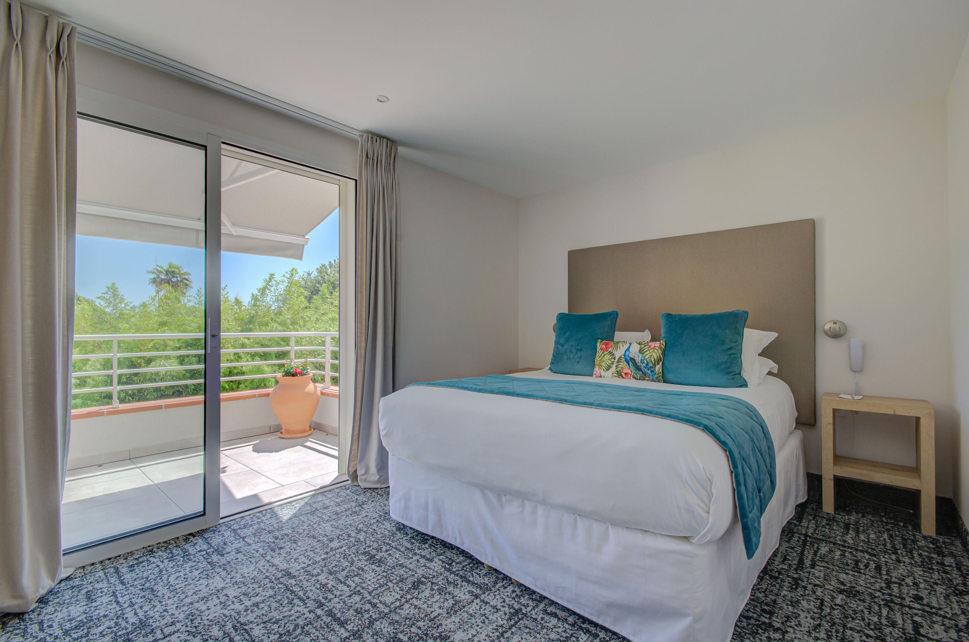 A hotel room with a large bed, a nightstand, a potted plant, and a sliding glass door leading to a balcony.