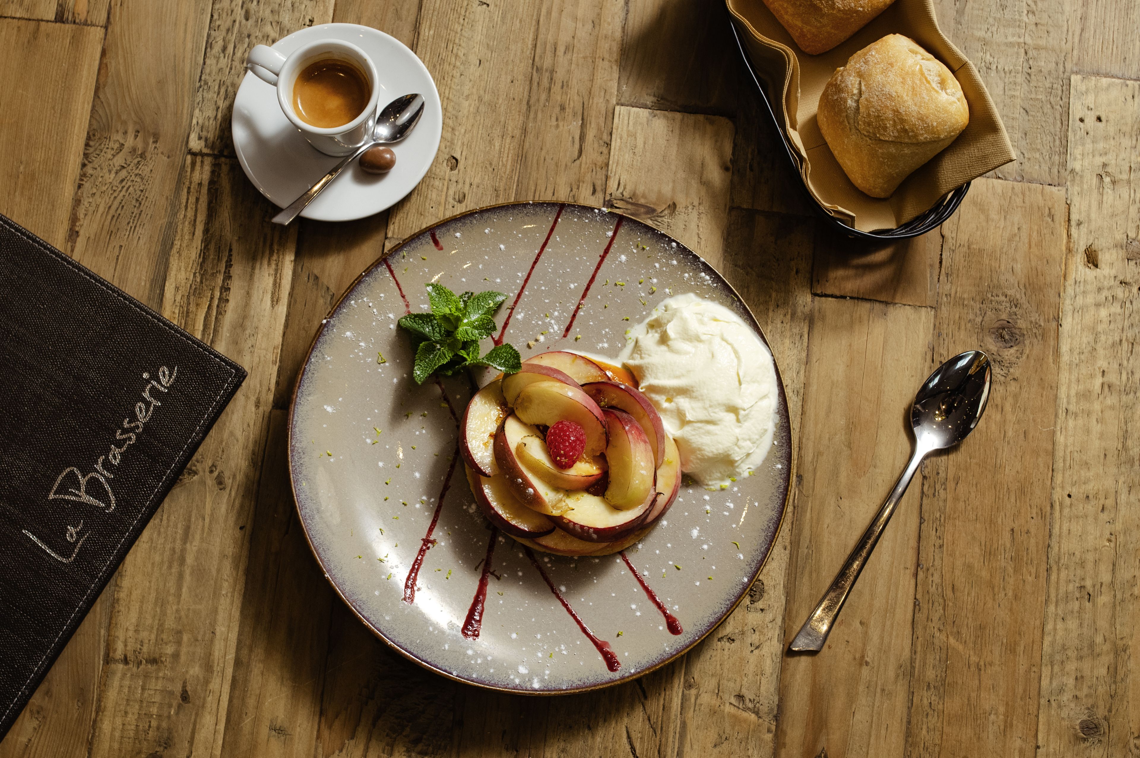 Une assiette de dessert avec des tranches de pomme, une fraise et de la crème fouettée, accompagnée d’une tasse de café et d’une cuillère sur une table en bois.