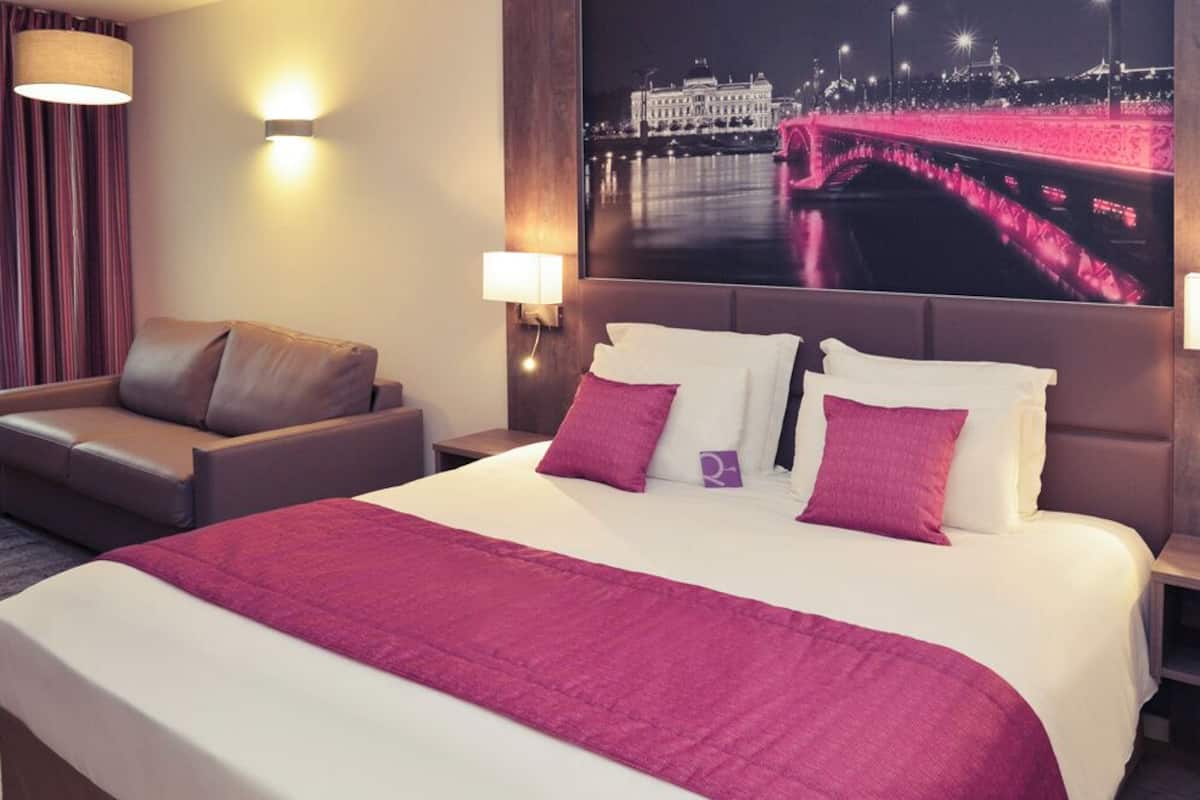 A hotel room with a large bed, two bedside tables, a chair, a wall-mounted TV displaying a cityscape, and a window with curtains.