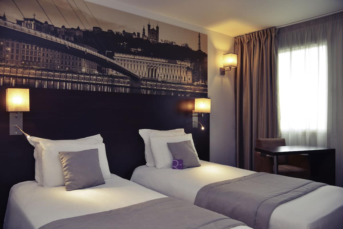 A hotel room with two beds, a cityscape mural on the wall, a desk, and a chair.
