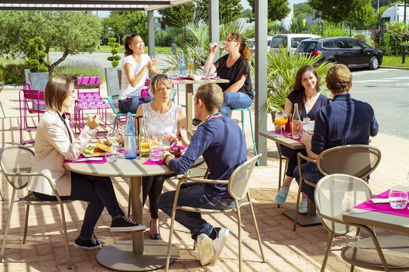A group of people dining outdoors at a cafe with tables and chairs, surrounded by greenery.