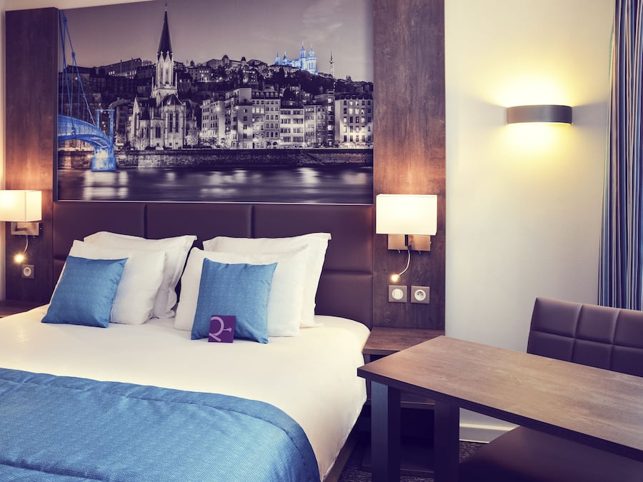 Et hotelværelse med en stor seng, to sengeborde, et skrivebord og en stol. Der er et bylandskabsmaleri på væggen bag sengen.