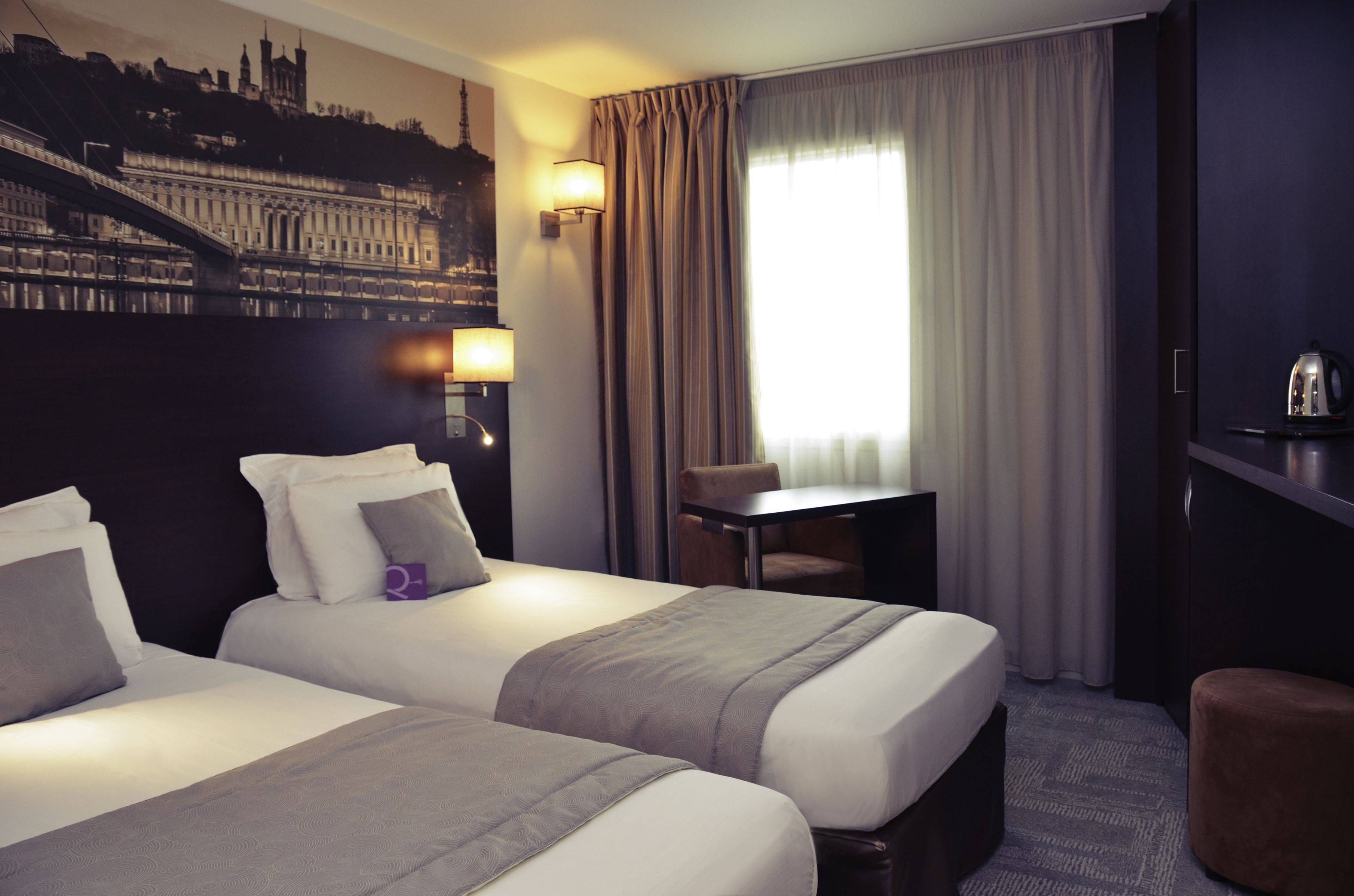 Quarto de hotel com duas camas, uma escrivaninha, uma cadeira, um abajur e vista para a cidade.