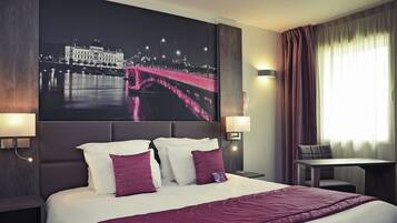 A hotel room with a large bed, two bedside lamps, a night view poster, a table, and a chair.