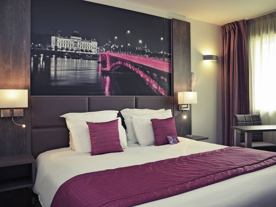 A hotel room with a large bed, two bedside lamps, a night view poster, a table, and a chair.