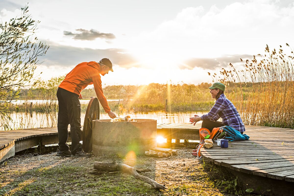 Two people by a campfire on a wooden dock with a lake in the background.
