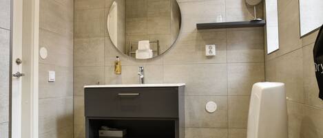 A modern bathroom with a round mirror, a toilet, a sink with a dark cabinet, and a trash can.