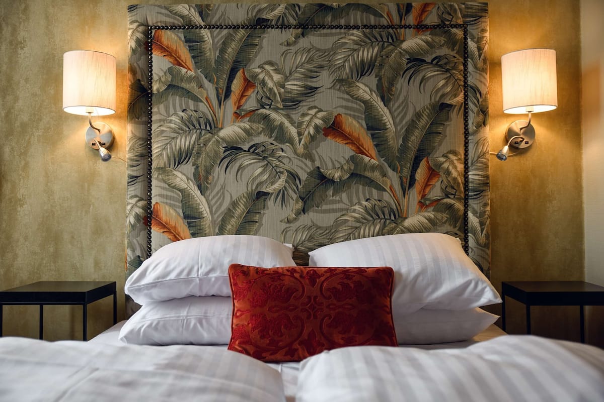 Une chambre d’hôtel avec une tête de lit ornée d’un motif de feuilles tropicales, deux lampes de chevet, du linge de lit blanc et un coussin rouge décoratif.